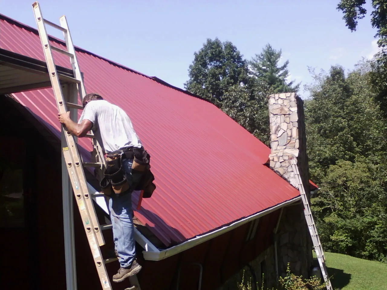 Roofer on a ladder beside a red metal roof and stone chimney — Soffit & Fascia Repair by Roofing Brothers in Chauncey